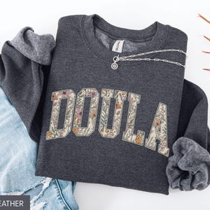 May include: Dark heather gray sweatshirt with the word "DOULA" in a floral design. The sweatshirt is folded with the sleeves showing a gray lining. A silver necklace is draped across the sweatshirt. Blue jeans are visible.