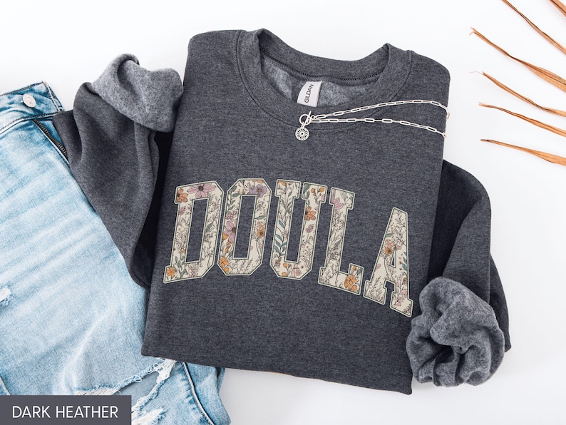 May include: Dark heather gray sweatshirt with the word "DOULA" in a floral design. The sweatshirt is folded with the sleeves showing a gray lining. A silver necklace is draped across the sweatshirt. Blue jeans are visible.