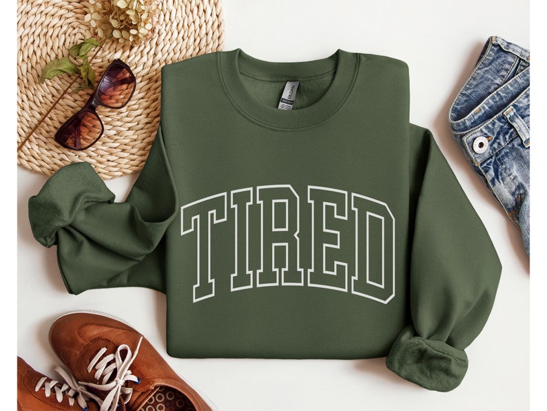 Pu&ograve; includere: Una felpa verde oliva con la scritta "TIRED" stampata in bianco, a blocchi. La felpa &egrave; esposta su una superficie bianca con scarpe marroni, occhiali da sole e un tappetino intrecciato.