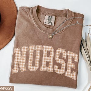 May include: Espresso-colored t-shirt with the word "NURSE" in a plaid pattern across the front. The shirt has a crew neck and short sleeves. A silver necklace with a small pendant is on the shirt. A brown hat is in the background.