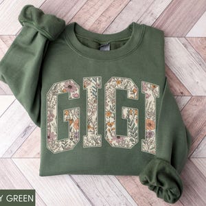 May include: Military green sweatshirt featuring the word "GIGI" in large, floral-filled letters. The letters are outlined in cream, and the flowers are in shades of pink, orange, and yellow. The sleeves are cuffed.