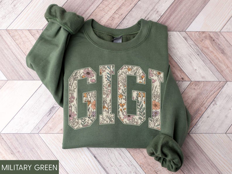 May include: Military green sweatshirt featuring the word "GIGI" in large, floral-filled letters. The letters are outlined in cream, and the flowers are in shades of pink, orange, and yellow. The sleeves are cuffed.