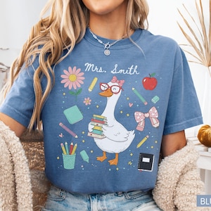 以下が含まれることがあります： 青いジーンズ色のTシャツ。気まぐれなグラフィックが特徴です。デザインは、メガネをかけ、本を運ぶガチョウ、学校をテーマにしたイラスト、そして「Mrs. Smith」という名前が特徴です。教師のためのキュートでカラフルなデザインです。