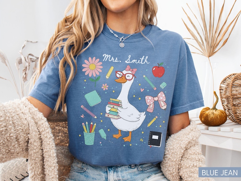 以下が含まれることがあります： 青いジーンズ色のTシャツ。気まぐれなグラフィックが特徴です。デザインは、メガネをかけ、本を運ぶガチョウ、学校をテーマにしたイラスト、そして「Mrs. Smith」という名前が特徴です。教師のためのキュートでカラフルなデザインです。