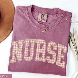 May include: Berry-colored t-shirt with the word "NURSE" in a plaid design. A gold necklace with a pendant rests on the shirt. A brown hat and dried plant are in the background. The shirt is a Comfort Colors brand.