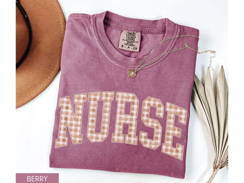 May include: Berry-colored t-shirt with the word "NURSE" in a plaid design. A gold necklace with a pendant rests on the shirt. A brown hat and dried plant are in the background. The shirt is a Comfort Colors brand.