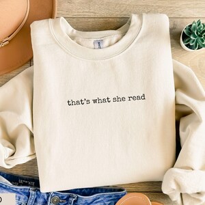 May include: A sand-colored sweatshirt with the phrase "that's what she read" in black lettering. The sweatshirt is folded on a wooden surface, with a brown hat, blue jeans, and a small succulent plant visible.