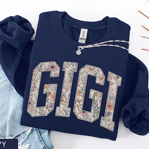 May include: A navy blue sweatshirt featuring the word "GIGI" in a floral design. A silver necklace with a pendant is draped across the sweatshirt. The sweatshirt is next to a pair of blue jeans.