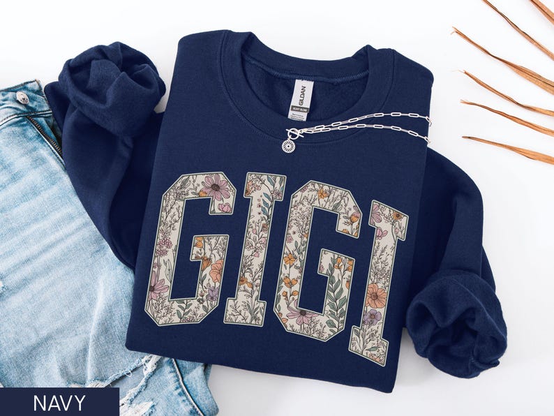 May include: A navy blue sweatshirt featuring the word "GIGI" in a floral design. A silver necklace with a pendant is draped across the sweatshirt. The sweatshirt is next to a pair of blue jeans.