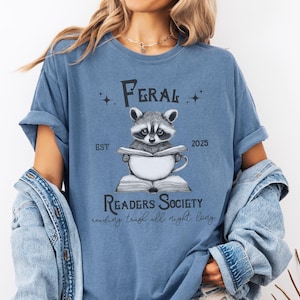 May include: A blue t-shirt with a graphic of a raccoon reading a book and drinking from a mug. The text on the shirt reads "Feral Readers Society est. 2025 reading trash all night long".