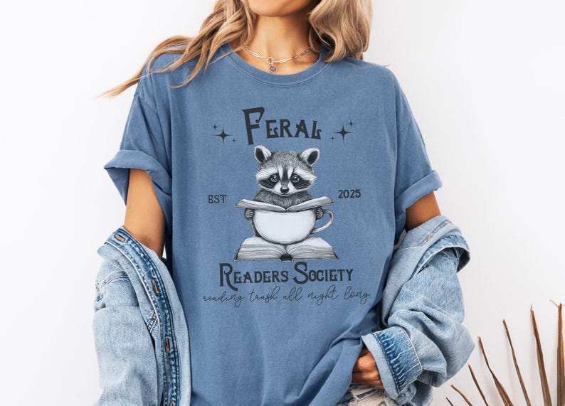May include: A blue t-shirt with a graphic of a raccoon reading a book and drinking from a mug. The text on the shirt reads "Feral Readers Society est. 2025 reading trash all night long".
