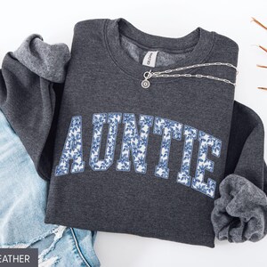 Peut inclure: Sweat-shirt gris fonc&eacute; avec le mot "AUNTIE" en grandes lettres fleuries bleues. Un collier en argent avec un pendentif est pos&eacute; sur le sweat-shirt. Les manches sont retrouss&eacute;es, et un jean bleu est visible.