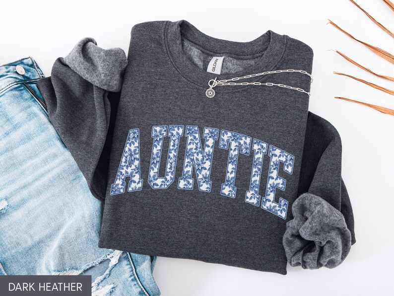 Peut inclure: Sweat-shirt gris fonc&eacute; avec le mot "AUNTIE" en grandes lettres fleuries bleues. Un collier en argent avec un pendentif est pos&eacute; sur le sweat-shirt. Les manches sont retrouss&eacute;es, et un jean bleu est visible.