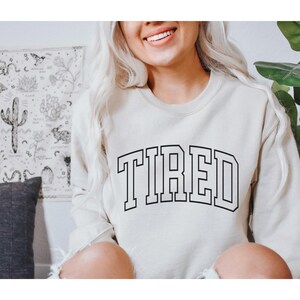 Pu&ograve; includere: Una felpa color crema con la scritta "TIRED" a grandi lettere nere. La felpa ha un classico girocollo e maniche lunghe. L'immagine mostra anche un arazzo decorativo e una pianta in vaso.