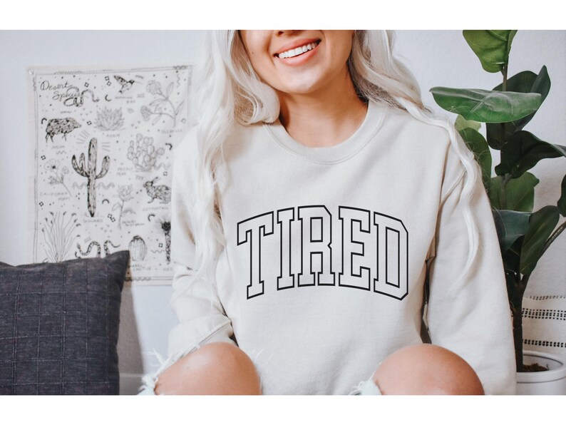 Pu&ograve; includere: Una felpa color crema con la scritta "TIRED" a grandi lettere nere. La felpa ha un classico girocollo e maniche lunghe. L'immagine mostra anche un arazzo decorativo e una pianta in vaso.
