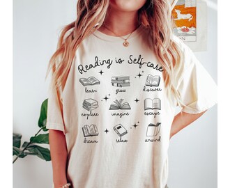 May include: A white t-shirt with the text "Reading is Self-care" and illustrations of books with words like "learn", "grow", "discover", "explore", "imagine", "escape", "dream", "relax", and "unwind" underneath each book.
