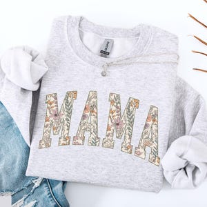 May include: A light gray sweatshirt with the word "MAMA" in a floral design across the front. The letters are filled with a pattern of pink, orange, and green flowers. A silver necklace with a small pendant rests on the sweatshirt.