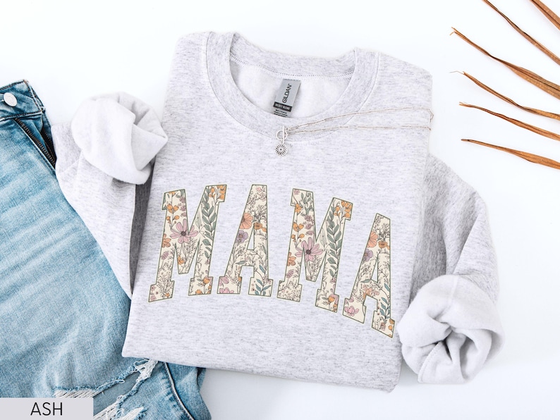 May include: A light gray sweatshirt with the word "MAMA" in a floral design across the front. The letters are filled with a pattern of pink, orange, and green flowers. A silver necklace with a small pendant rests on the sweatshirt.