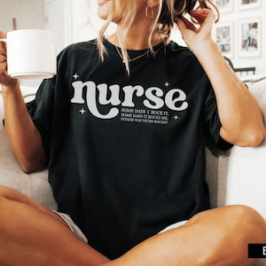 Pode incluir: T-shirt preta com a palavra "nurse" em fonte retr&ocirc; branca, com o texto "SOME DAYS I ROCK IT, SOME DAYS IT ROCKS ME, EITHER WAY WE'RE ROCKIN" abaixo. A camisa &eacute; de estilo casual.