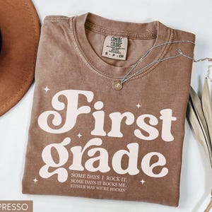 Puede incluir: Camiseta color espresso con las palabras "First Grade" en una fuente retro. La camiseta tambi&eacute;n presenta el texto "SOME DAYS I ROCK IT, SOME DAYS IT ROCKS ME, EITHER WAY WE'RE ROCKIN'". Tambi&eacute;n se ve un collar plateado y un sombrero marr&oacute;n.
