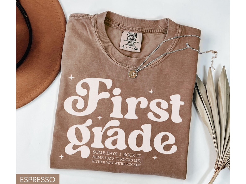 Puede incluir: Camiseta color espresso con las palabras "First Grade" en una fuente retro. La camiseta tambi&eacute;n presenta el texto "SOME DAYS I ROCK IT, SOME DAYS IT ROCKS ME, EITHER WAY WE'RE ROCKIN'". Tambi&eacute;n se ve un collar plateado y un sombrero marr&oacute;n.