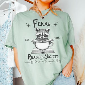 May include: A light green t-shirt with a graphic of a raccoon reading a book in a mug. The text on the shirt reads "Feral Readers Society est 2025 reading trash all night long".
