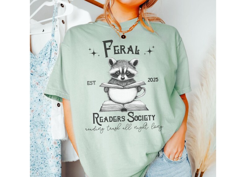 May include: A light green t-shirt with a graphic of a raccoon reading a book in a mug. The text on the shirt reads "Feral Readers Society est 2025 reading trash all night long".