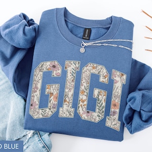 May include: Indigo blue sweatshirt featuring the name "GIGI" in a floral pattern. The sweatshirt is folded on blue denim jeans. A silver necklace with a small pendant is on the sweatshirt.