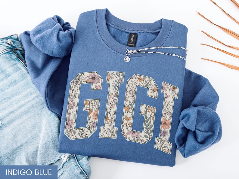 May include: Indigo blue sweatshirt featuring the name "GIGI" in a floral pattern. The sweatshirt is folded on blue denim jeans. A silver necklace with a small pendant is on the sweatshirt.