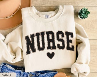 May include: A sand-colored sweatshirt with the word "NURSE" in large, black, leopard-print letters. A black heart is below the word. The sweatshirt is folded on a wooden surface, with a hat and jeans visible.