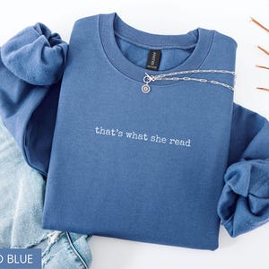 May include: Indigo blue sweatshirt with the embroidered text "that's what she read." A silver necklace with a pendant is draped across the front. The sweatshirt is folded on top of a pair of blue jeans.