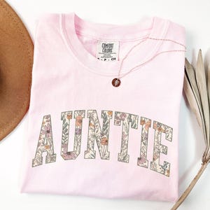 May include: Pink t-shirt with a floral print design that says "AUNTIE" in a varsity style font.