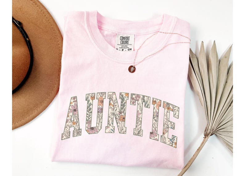 May include: Pink t-shirt with a floral print design that says "AUNTIE" in a varsity style font.