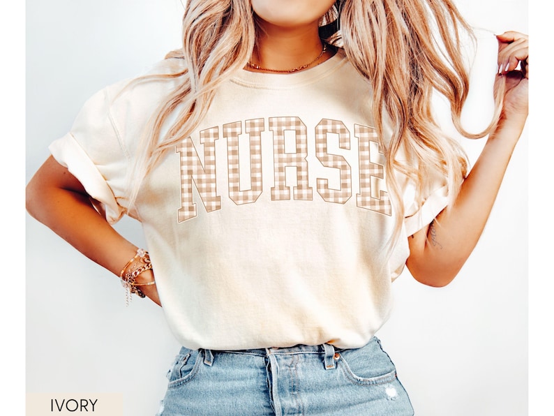 May include: Ivory-colored t-shirt with the word "NURSE" in a tan and white plaid design across the chest. The shirt has short sleeves and a relaxed fit. The word "IVORY" is printed at the bottom of the image.