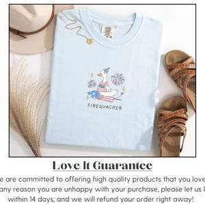 Op de afbeelding: Lichtblauw T-shirt met een cartoonillustratie van een gans die een feestmuts draagt en op een vuurwerk rijdt. De tekst "FIREQUACKER" is onder de illustratie gedrukt. Het T-shirt ligt op een wit oppervlak met een strohoed en sandalen.