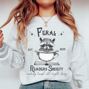 May include: A gray sweatshirt with the text "Feral Readers Society" and a raccoon holding a book and a mug. The text below the raccoon says "reading trash all night long".