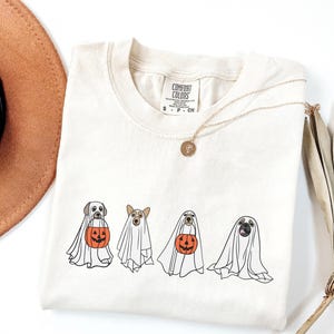 Puede incluir: Camiseta color crema con cuatro perros disfrazados de fantasmas, cada uno con una apariencia espec&iacute;fica de raza. Los perros sostienen calabazas de Halloween naranjas. Tambi&eacute;n se ve un collar dorado y un sombrero marr&oacute;n.