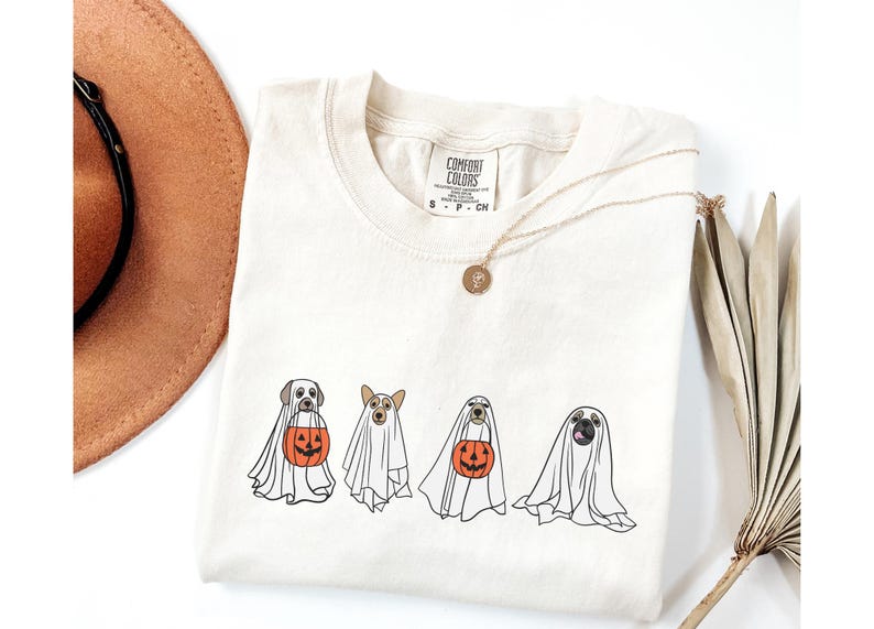 Puede incluir: Camiseta color crema con cuatro perros disfrazados de fantasmas, cada uno con una apariencia espec&iacute;fica de raza. Los perros sostienen calabazas de Halloween naranjas. Tambi&eacute;n se ve un collar dorado y un sombrero marr&oacute;n.