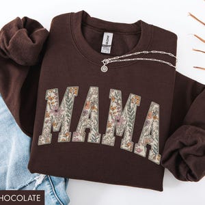 May include: Dark chocolate brown sweatshirt with the word "MAMA" in a floral design. A silver necklace with a pendant is draped across the sweatshirt. The sweatshirt is folded next to a pair of blue jeans.