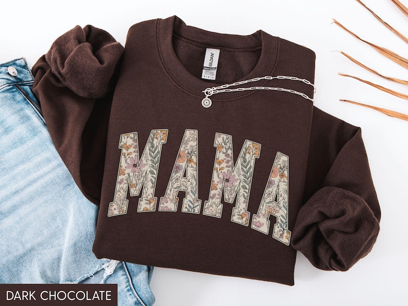 May include: Dark chocolate brown sweatshirt with the word "MAMA" in a floral design. A silver necklace with a pendant is draped across the sweatshirt. The sweatshirt is folded next to a pair of blue jeans.