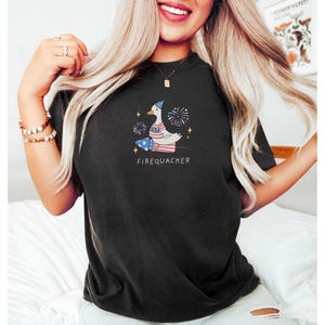 Op de afbeelding: Zwart T-shirt met een witte eend die een rood-wit-blauwe hoed draagt en een vuurwerk vasthoudt. De eend is omringd door sterren en vuurwerk. De tekst "FIREQUACKER" is onder de eend gedrukt.