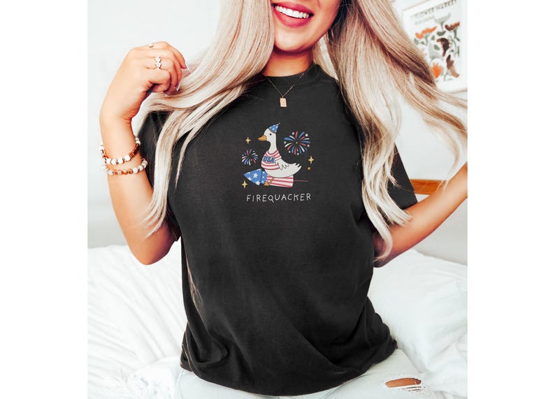 Op de afbeelding: Zwart T-shirt met een witte eend die een rood-wit-blauwe hoed draagt en een vuurwerk vasthoudt. De eend is omringd door sterren en vuurwerk. De tekst "FIREQUACKER" is onder de eend gedrukt.
