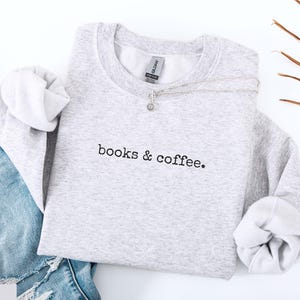 May include: A light gray sweatshirt with the text "books & coffee." is displayed on a white surface next to a pair of blue jeans. A silver necklace with a small pendant rests on the sweatshirt.