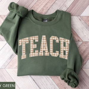 May include: Military green sweatshirt with the word "TEACH" in large, tan and white gingham letters across the chest. The sweatshirt has long sleeves and a crew neck. The text "MILITARY GREEN" is at the bottom.