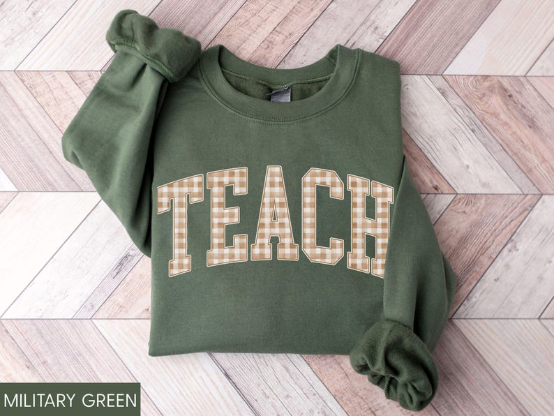 May include: Military green sweatshirt with the word "TEACH" in large, tan and white gingham letters across the chest. The sweatshirt has long sleeves and a crew neck. The text "MILITARY GREEN" is at the bottom.