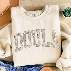 May include: Cream-colored sweatshirt with the word "DOULA" in large, floral-filled letters. The letters are outlined in a darker color, and the floral design includes shades of purple, orange, and green. The sweatshirt is folded on a wooden surface, with a hat and jeans visible.
