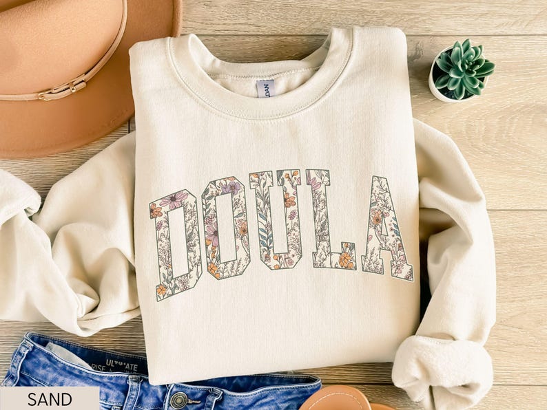 May include: Cream-colored sweatshirt with the word "DOULA" in large, floral-filled letters. The letters are outlined in a darker color, and the floral design includes shades of purple, orange, and green. The sweatshirt is folded on a wooden surface, with a hat and jeans visible.
