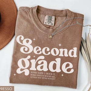 May include: Espresso-colored t-shirt with the words "Second Grade" in a retro font. The shirt also has the text "Some days I rock it, some days it rocks me, either way we're rockin". A silver necklace with a pendant is on the shirt.