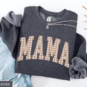 May include: Dark heather gray sweatshirt with the word "MAMA" in a tan and white gingham pattern across the front. A silver necklace is draped across the sweatshirt. The sweatshirt is folded next to a pair of blue jeans.