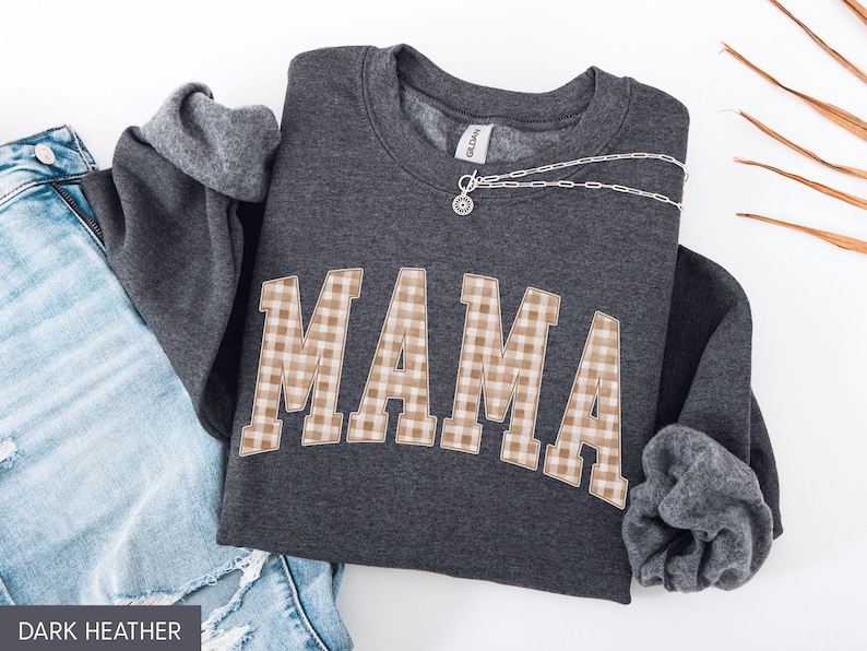May include: Dark heather gray sweatshirt with the word "MAMA" in a tan and white gingham pattern across the front. A silver necklace is draped across the sweatshirt. The sweatshirt is folded next to a pair of blue jeans.
